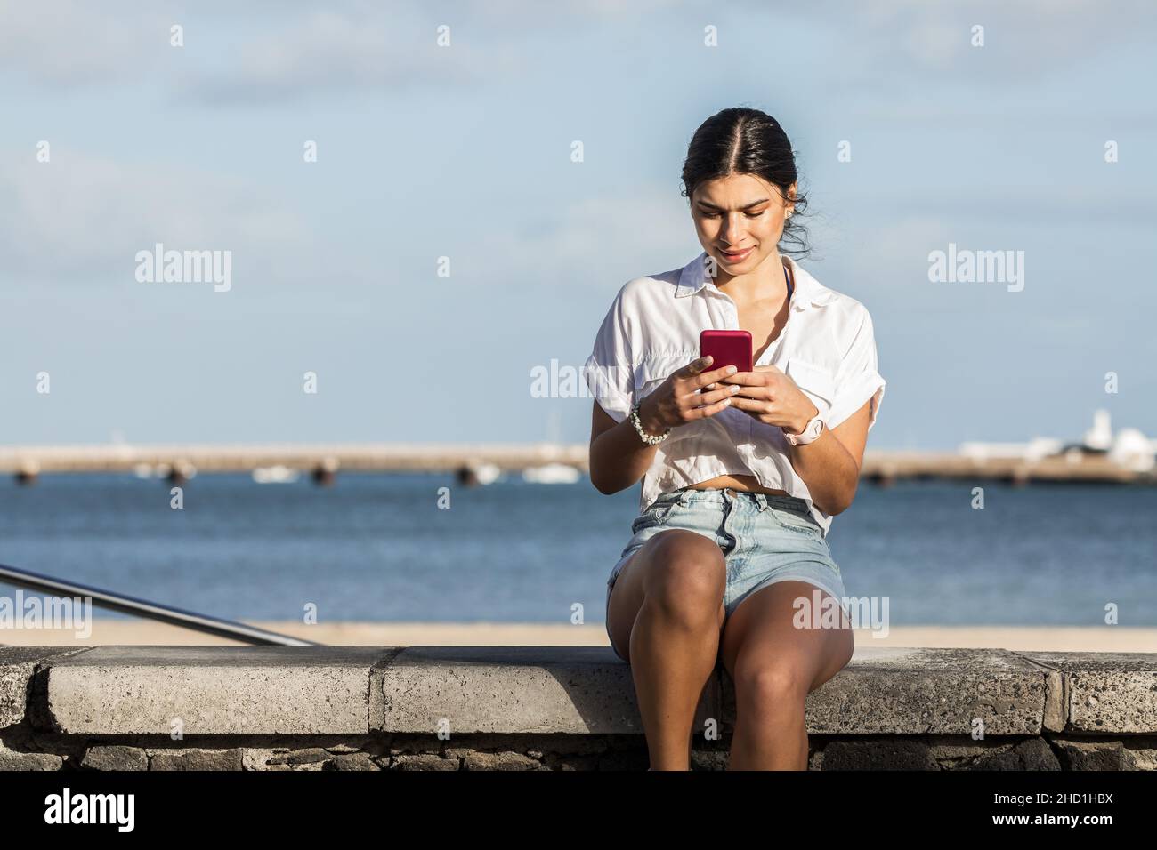 Charmante femme en tenue d'été assise sur la clôture sur fond de rivage sur Lanzarote et surfer sur Internet sur smartphone tout en appréciant le week-end Banque D'Images