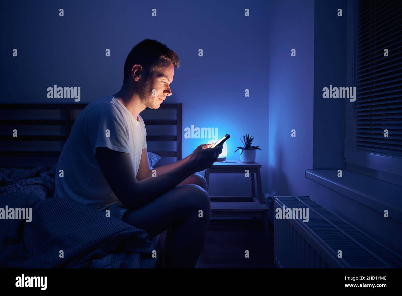 Homme solitaire assis sur le lit dans la chambre et utilisant le téléphone la nuit.Visage éclairé depuis l'écran du smartphone. Banque D'Images