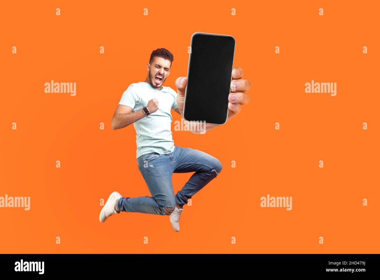 Homme heureux volant et sautant dans l'air et montrant grand écran mobile vide pour l'espace de copie et la zone publicitaire. Studio d'intérieur tourné isolé sur fond orange Banque D'Images