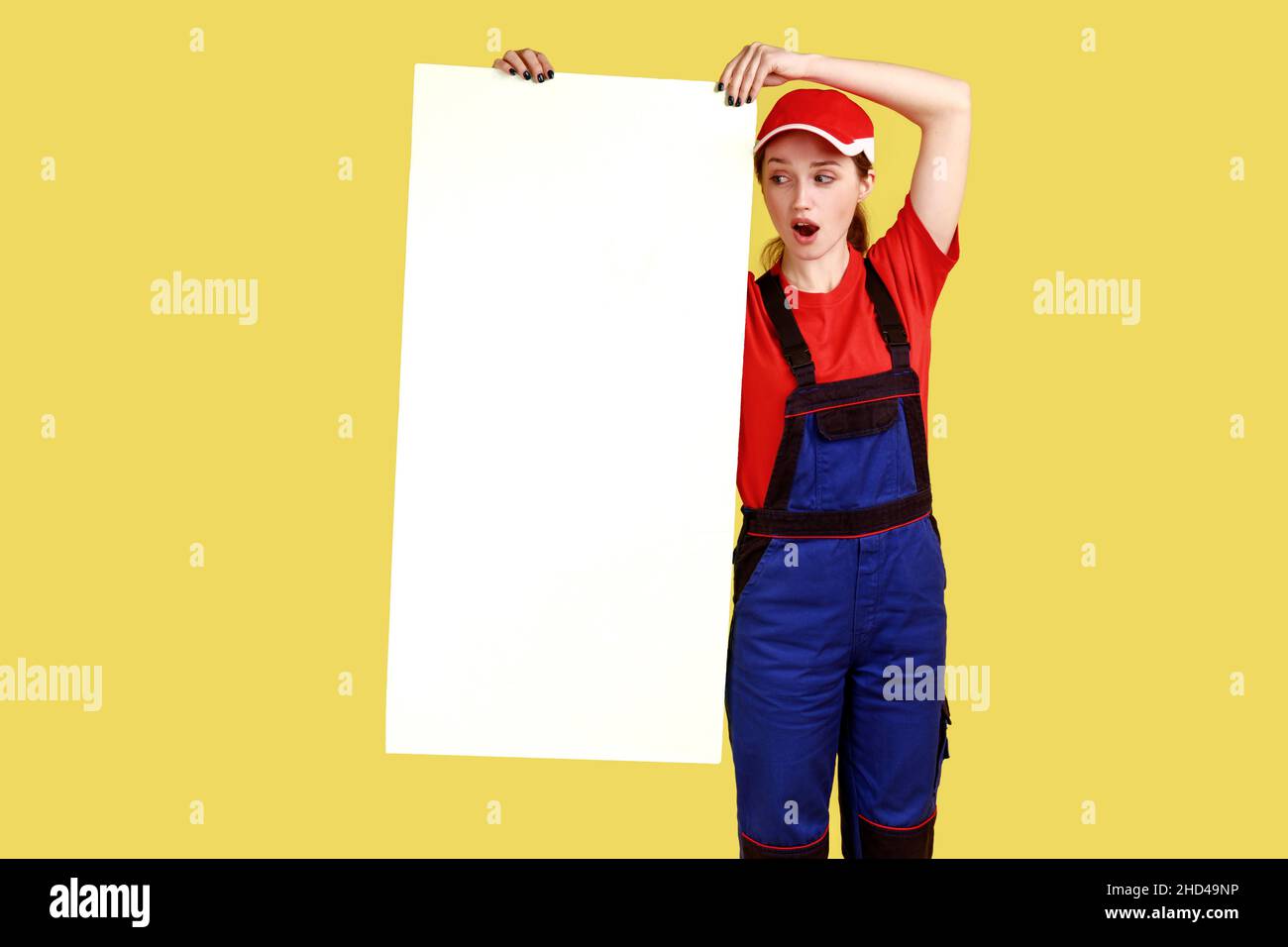 Portrait en longueur de femme sous le choc architecte debout et montrant une grande affiche blanche avec espace vide pour la publicité, portant une combinaison et une casquette.Studio d'intérieur isolé sur fond jaune. Banque D'Images