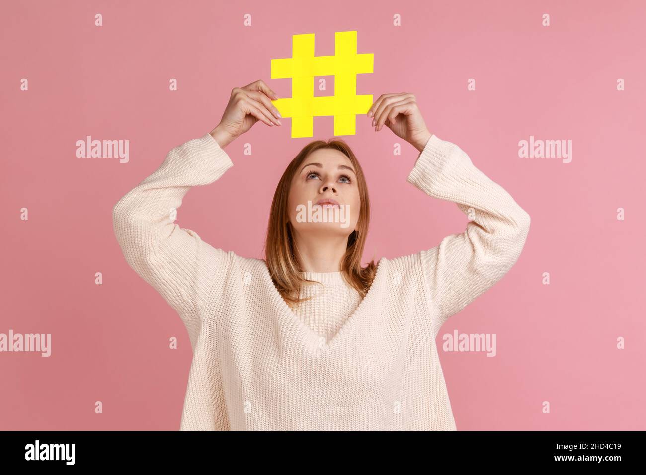 Portrait de belle femme blonde tenant le signe hashtag au-dessus de la tête, poste populaire dans Internet, recherche facile vers le haut, portant chandail blanc.Studio d'intérieur isolé sur fond rose. Banque D'Images