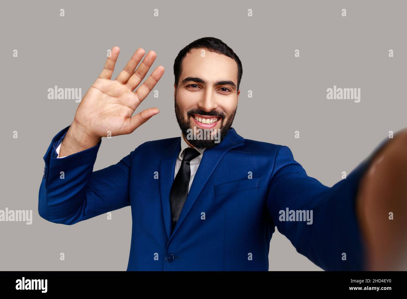 Homme d'affaires positif sympathique barbu faisant selfie ou streaming, en agitant main POV, point de vue de la photo, portant le costume de style officiel.Prise de vue en studio isolée sur fond gris. Banque D'Images