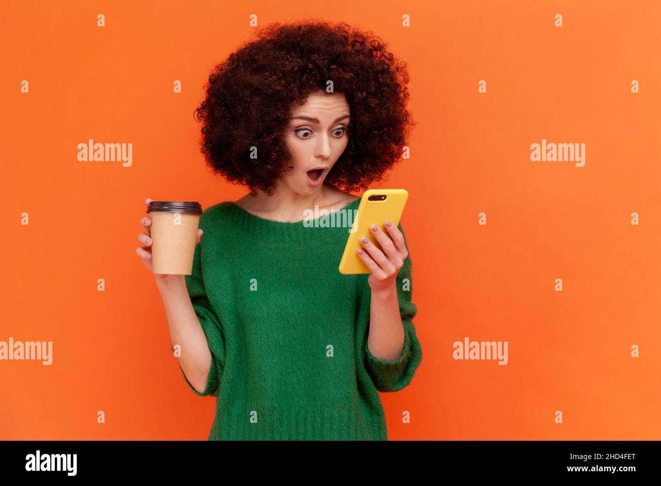 Femme stupéfiante avec une coiffure afro en vert chandail décontracté debout avec un smartphone dans les mains, boire un café à emporter, lire des nouvelles choquantes.Studio d'intérieur isolé sur fond orange. Banque D'Images