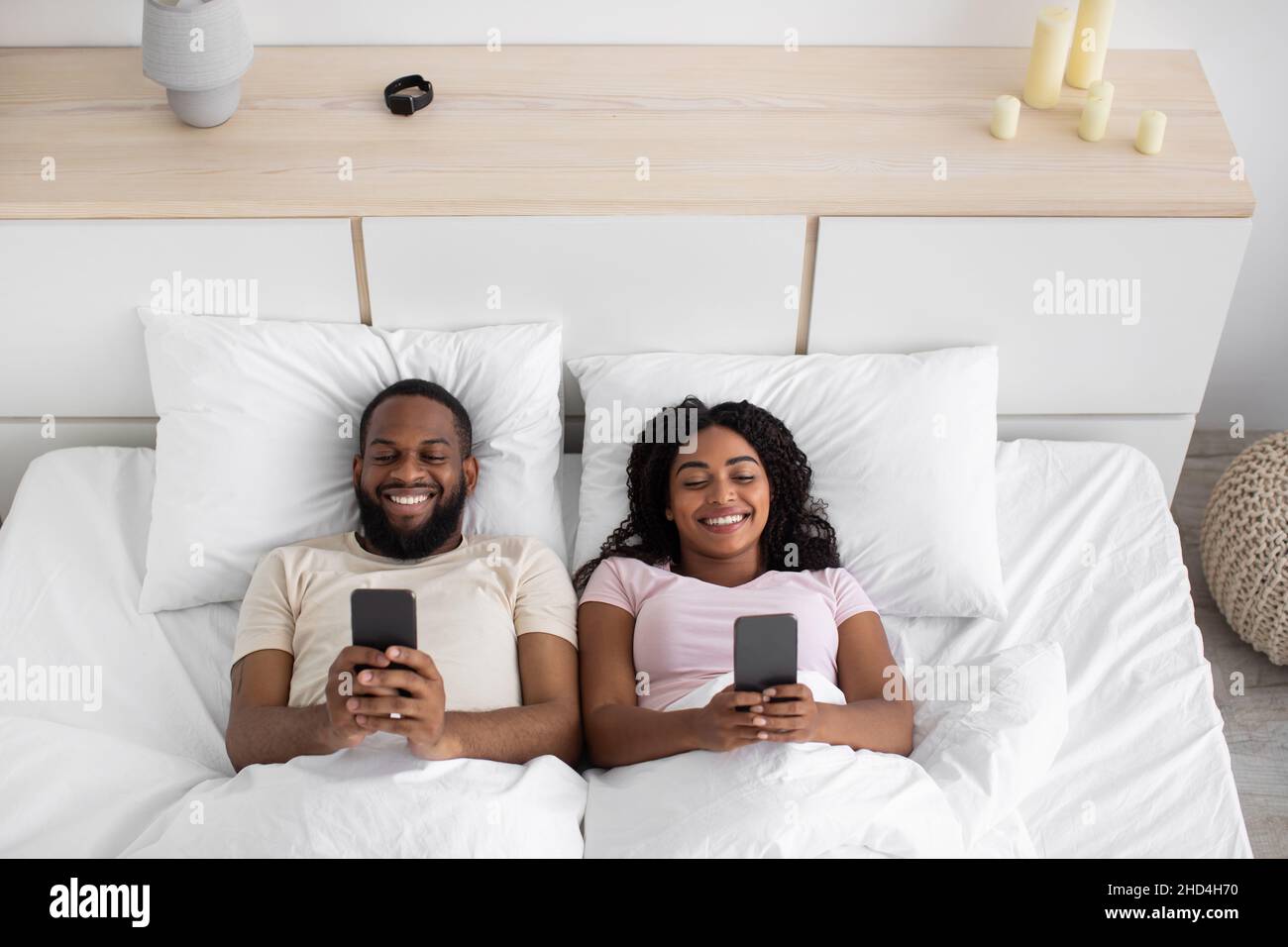 Souriant jeune afro-américain mari et femme dactylographiant sur les téléphones, jouant en ligne jeux sur le lit dans la chambre à l'intérieur Banque D'Images