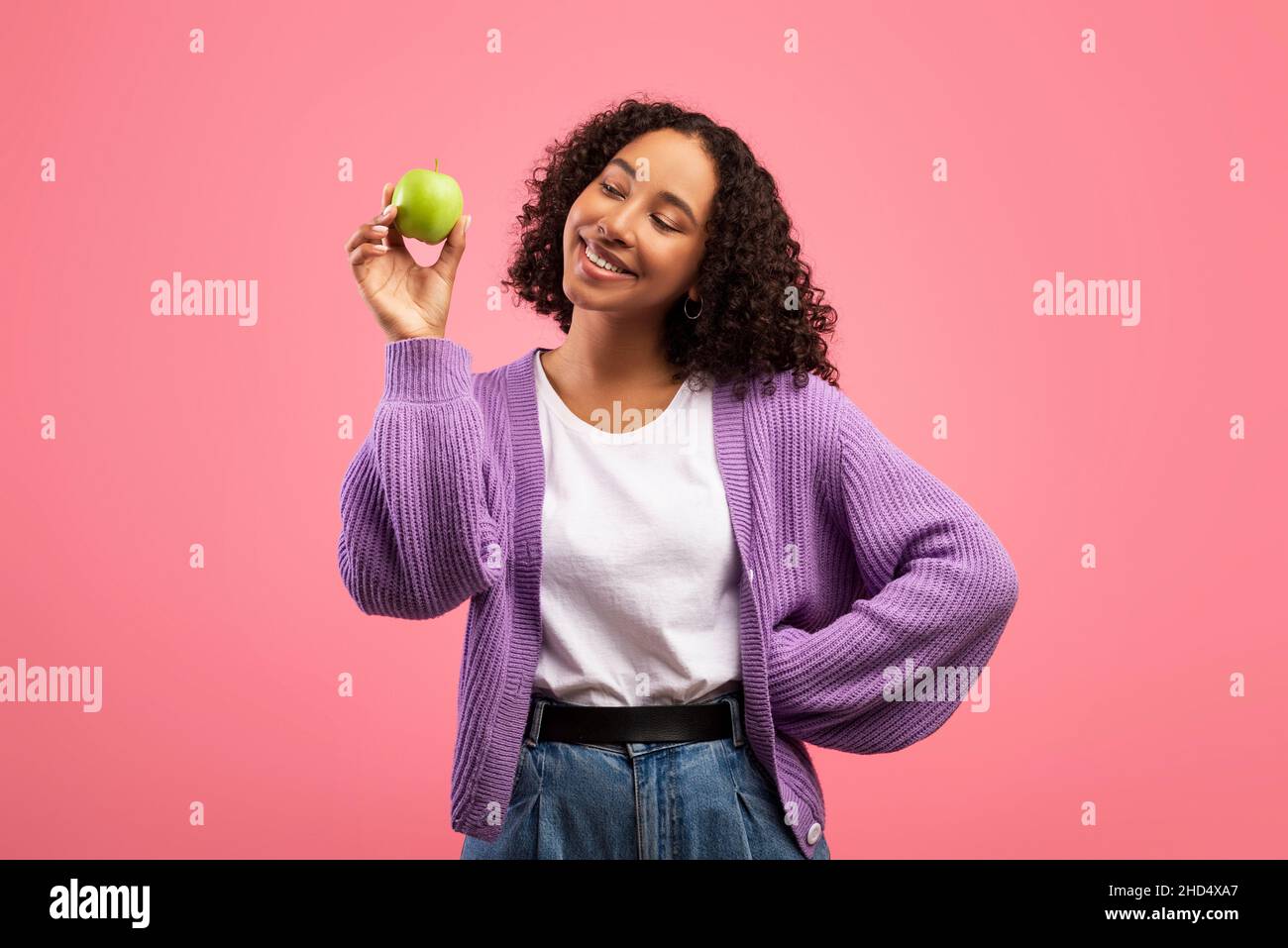 Portrait de jeune femme noire gaie tenant une pomme verte mûre et souriant sur fond rose studio Banque D'Images
