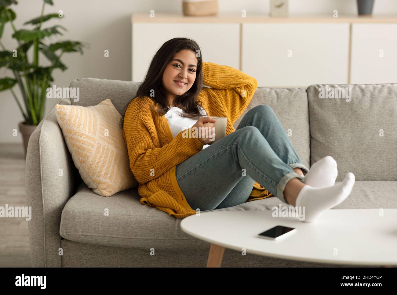 Jolie femme arabe qui se rafraîchissante sur un canapé avec une tasse de boisson chaude à la maison, passant un week-end paresseux avec un café aromatique Banque D'Images