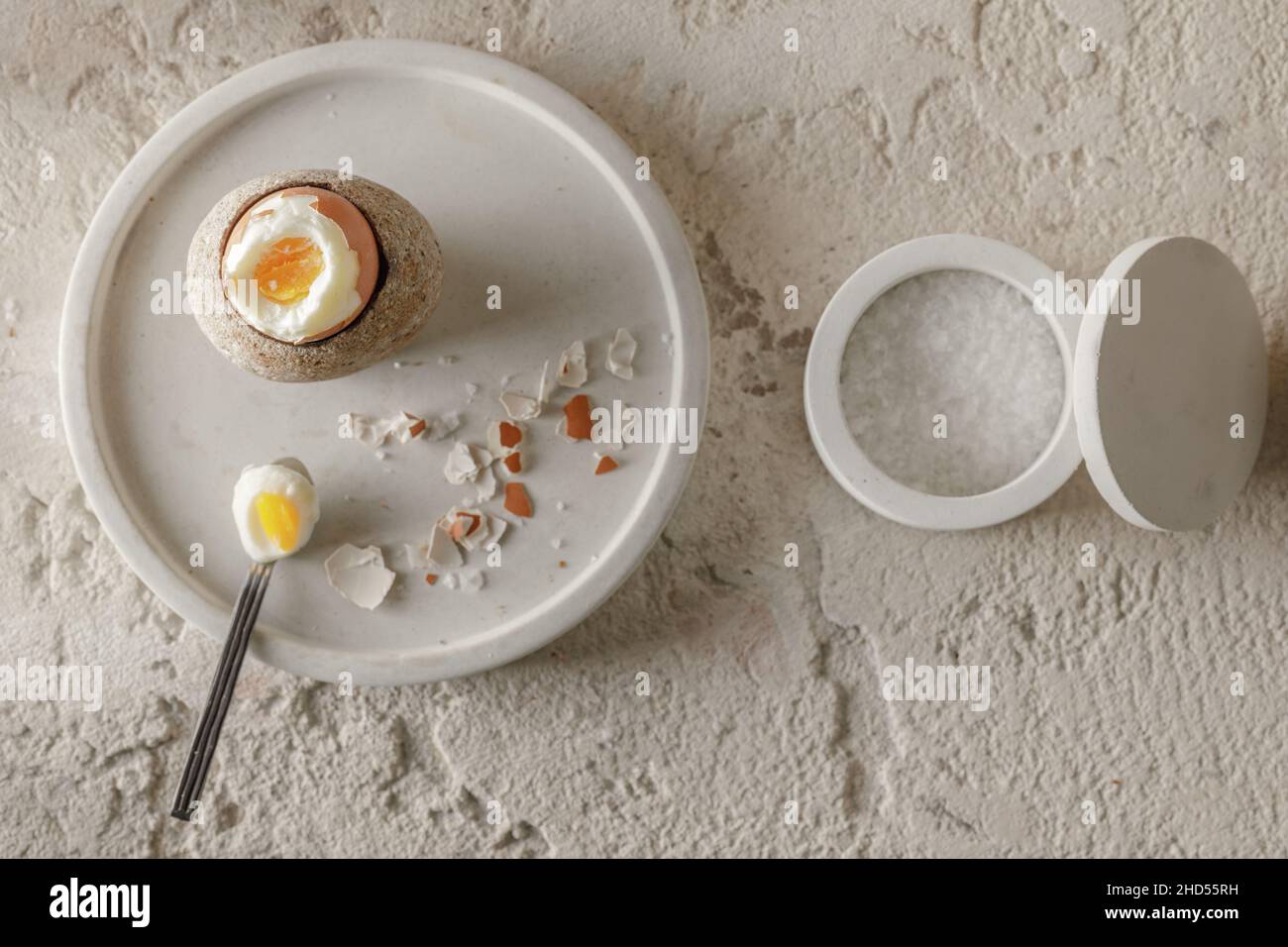 Un œuf dur dans un coquetier en grès avec morceaux de coquetier cassés dans un plateau en béton, flocons de sel dans des bols en béton blanc sur un backgrou en argile rugueux Banque D'Images