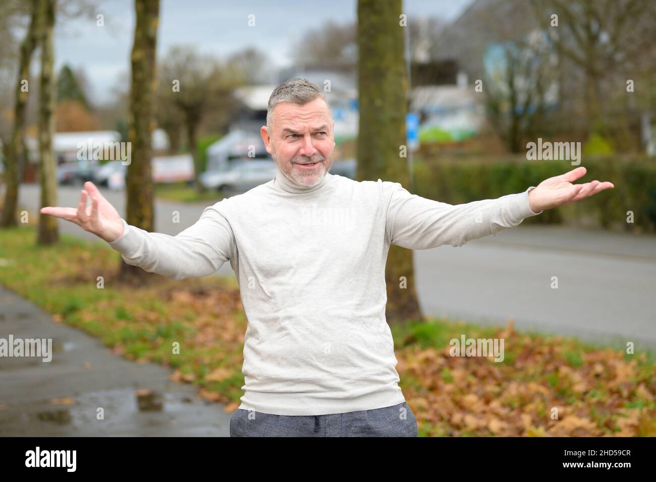 Homme âgé faisant un geste expansif avec ses bras les étirant large avec un sourire tranquille à l'extérieur dans une rue urbaine Banque D'Images