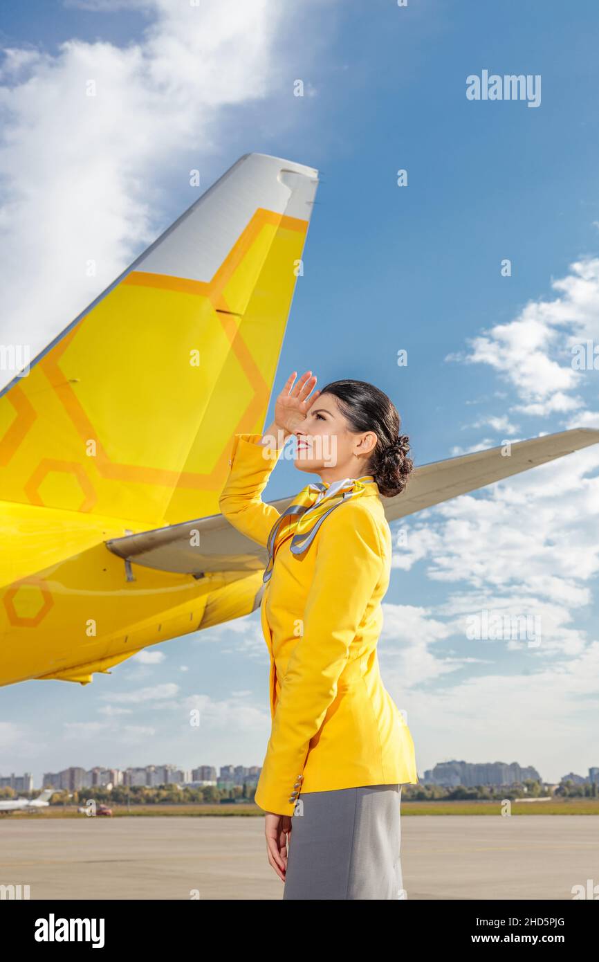 Une hôtesse de vol gaie couvrant les yeux du soleil avec sa main et souriant tout en se tenant près de l'avion jaune à l'aéroport Banque D'Images