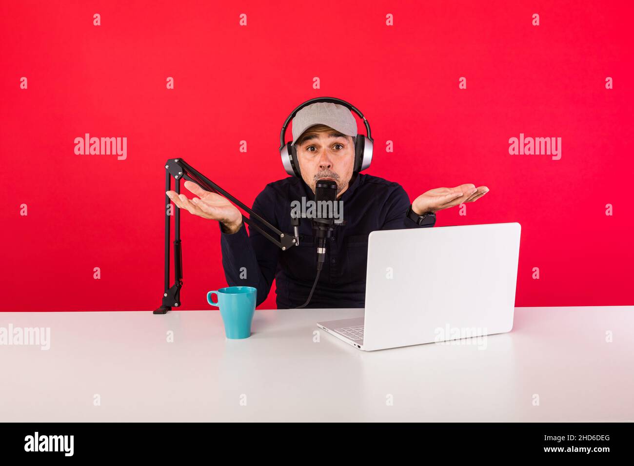 Homme présentateur avec Cap dans le studio d'enregistrement radio de podcast faisant des gestes de main, à côté d'un ordinateur et d'un microphone , sur fond rouge.Podcasting, br Banque D'Images