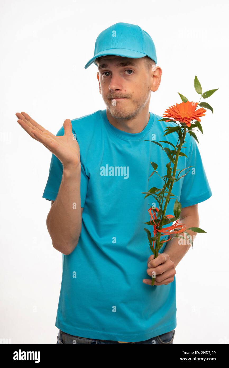 Jeune beau grand homme blanc mince avec des cheveux bruns regardant négligent tenant fleur orange avec chemise bleue et capuchon bleu isolé sur fond blanc Banque D'Images