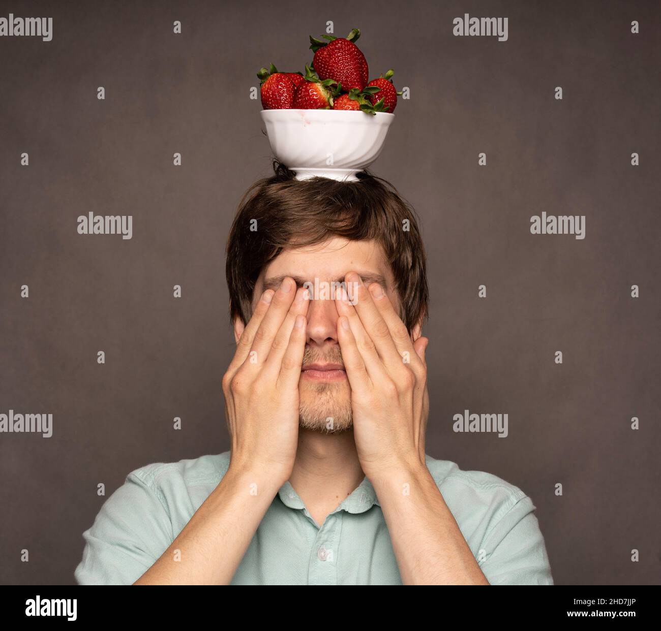 Jeune homme blanc mince et élégant, avec un bol d'équilibre de cheveux bruns de fraises sur la tête couvrant ses yeux sur fond gris Banque D'Images