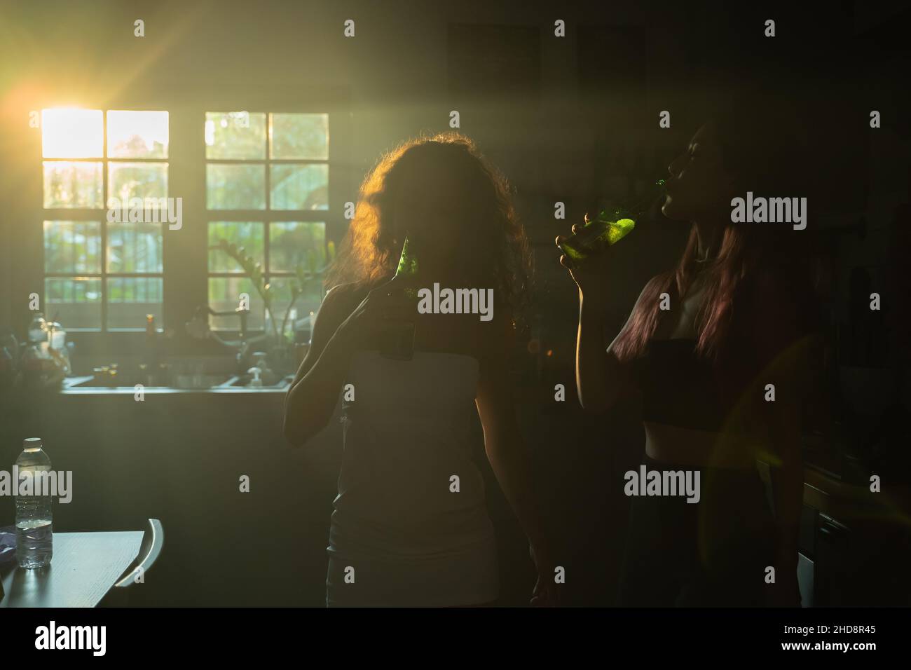 Groupe de deux jeunes amies qui boivent de la bière à la bouteille après une soirée à la maison au lever du soleil en se tenant dans la cuisine Banque D'Images