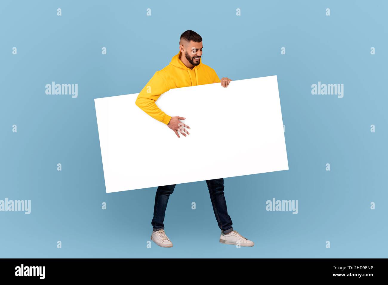 Homme arabe excité marchant avec un écriteau blanc sous le bras sur fond bleu de studio, maquette pour le texte ou le design Banque D'Images