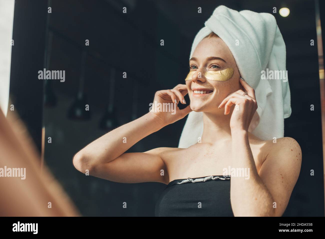 Gros plan portrait d'une fille à cheveux rouges avec une serviette sur la tête en appliquant des taches oculaires.Femme caucasienne souriante.Soin du visage.Nettoyer la peau.Beauté Banque D'Images