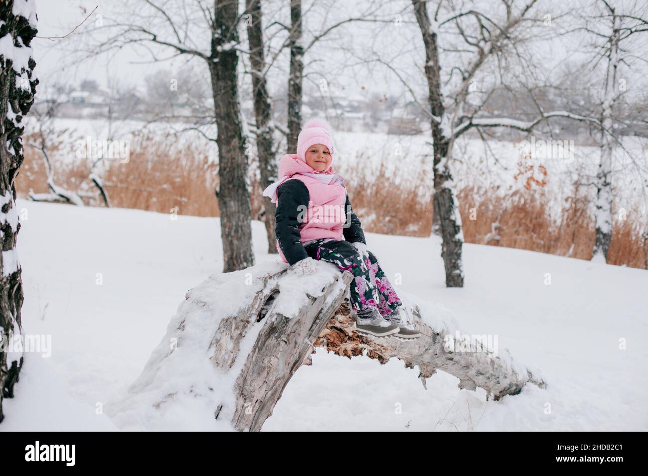 Portrait éloigné de la femme russe enfant regardant dans l'appareil photo assis sur un arbre et souriant portant des vêtements d'hiver roses dans la forêt.Étonnant Banque D'Images