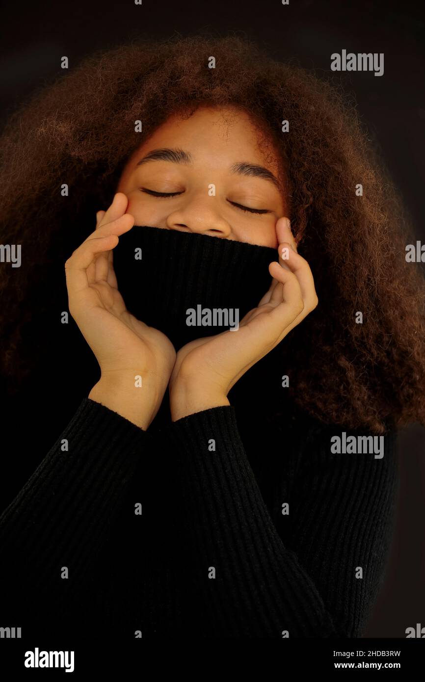 Heureuse afro-américaine curly femme fermer les yeux et couvrant le visage avec chaud noir col pull isolé sur fond sombre studio, souriante femme Banque D'Images
