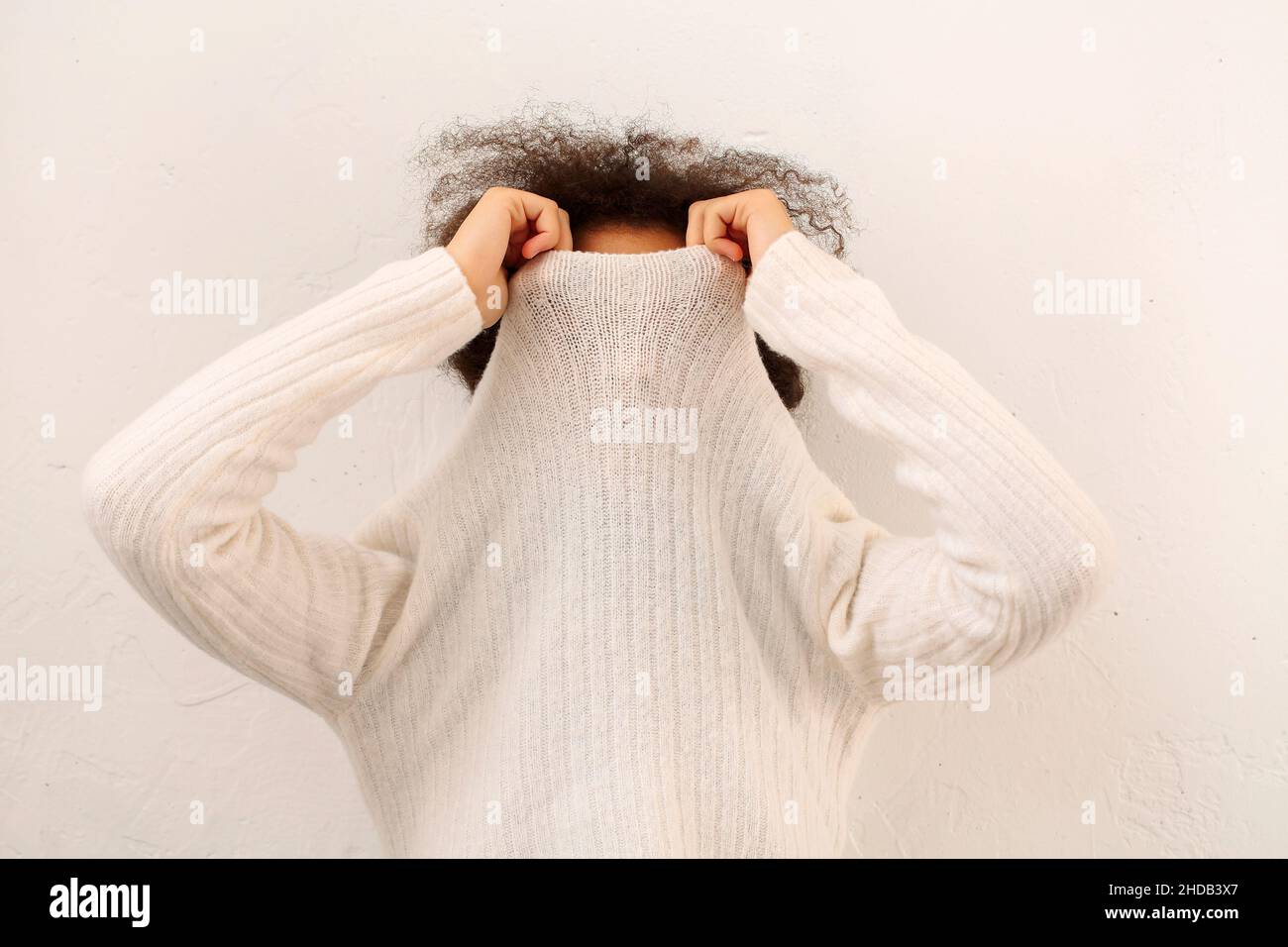 Une femme en forme de curly africaine couvre la face avec un col chandail blanc chaud isolé sur fond studio léger, une femme qui prend le pull à col roulé, ne peut pas chauffer Banque D'Images