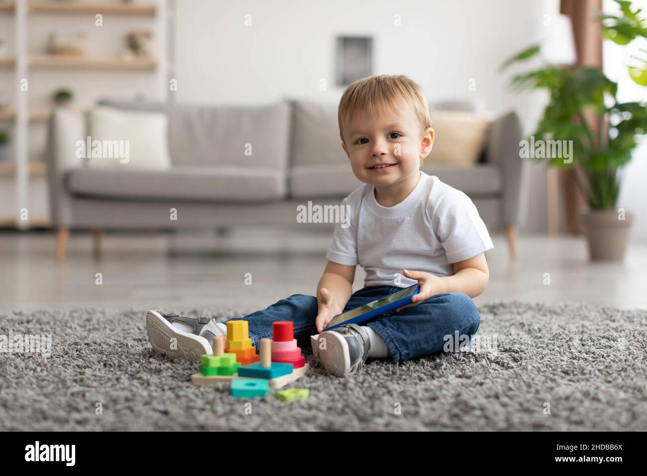 Un petit garçon heureux regardant un dessin animé sur un téléphone intelligent tout en étant assis sur la moquette, en regardant et en souriant l'appareil photo Banque D'Images