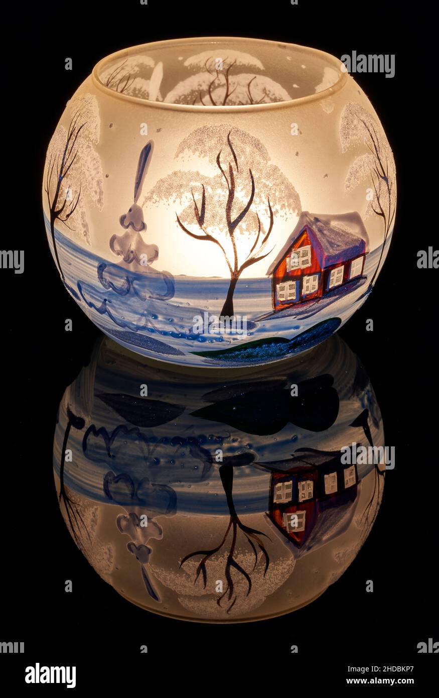Une tasse en verre peinte à la main avec un décor hivernal éclairé par une bougie à l'intérieur de la tasse.Un gros plan photographique utilisant la peinture légère. Banque D'Images