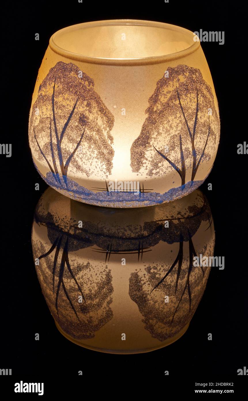 Une tasse en verre peinte à la main avec un décor hivernal éclairé par une bougie à l'intérieur de la tasse.Un gros plan photographique utilisant la peinture légère. Banque D'Images