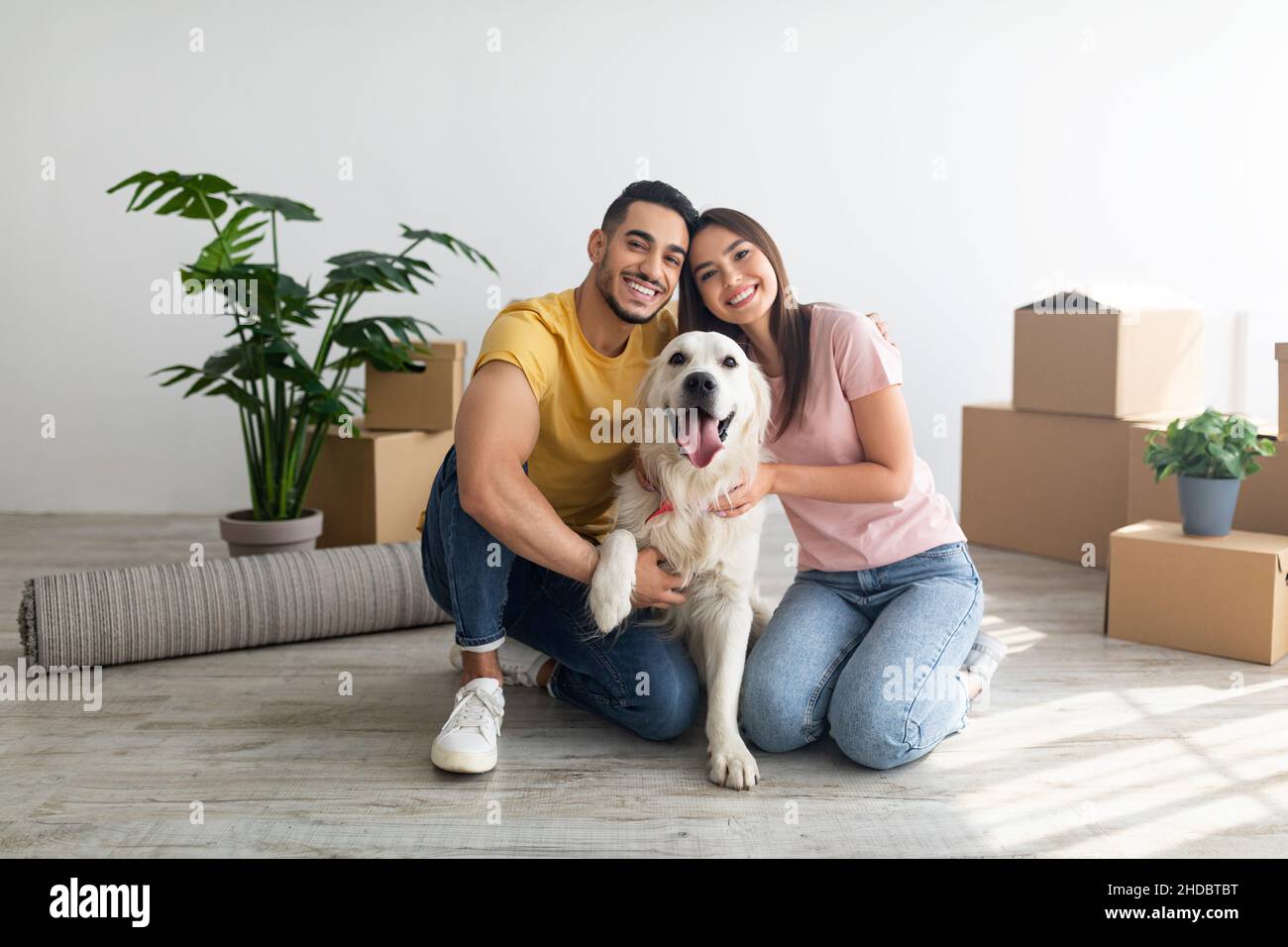 Jeune couple international gaie avec un adorable chien Golden Retriever assis au sol de la nouvelle maison le jour de la réinstallation Banque D'Images