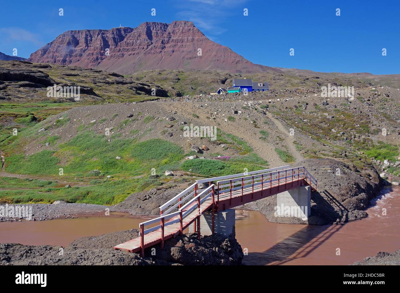 Rivière brun rougeâtre traversant un paysage arctique vert, pont en bois simple et huttes, île de Disko, Qeqertarsuaq, Arctique, Groenland,Danemark, N Banque D'Images