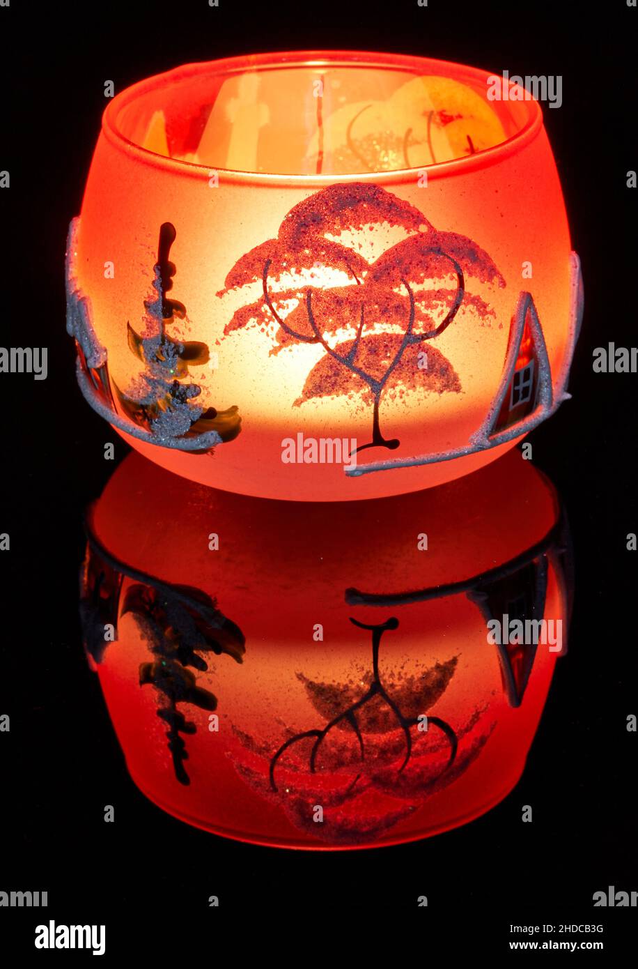 Une tasse en verre peinte à la main avec un décor hivernal éclairé par une bougie à l'intérieur de la tasse.Un gros plan photographique utilisant la peinture légère. Banque D'Images