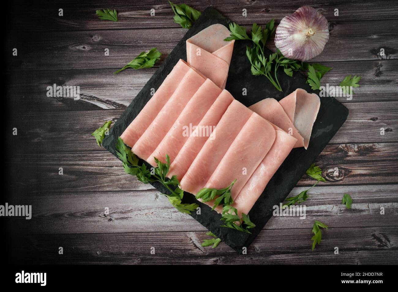 Vue en hauteur d'un arrangement élégant sur une dalle noire de jambon cuit ou prosciutto cotto avec un ail sur un panneau rustique en bois. Banque D'Images