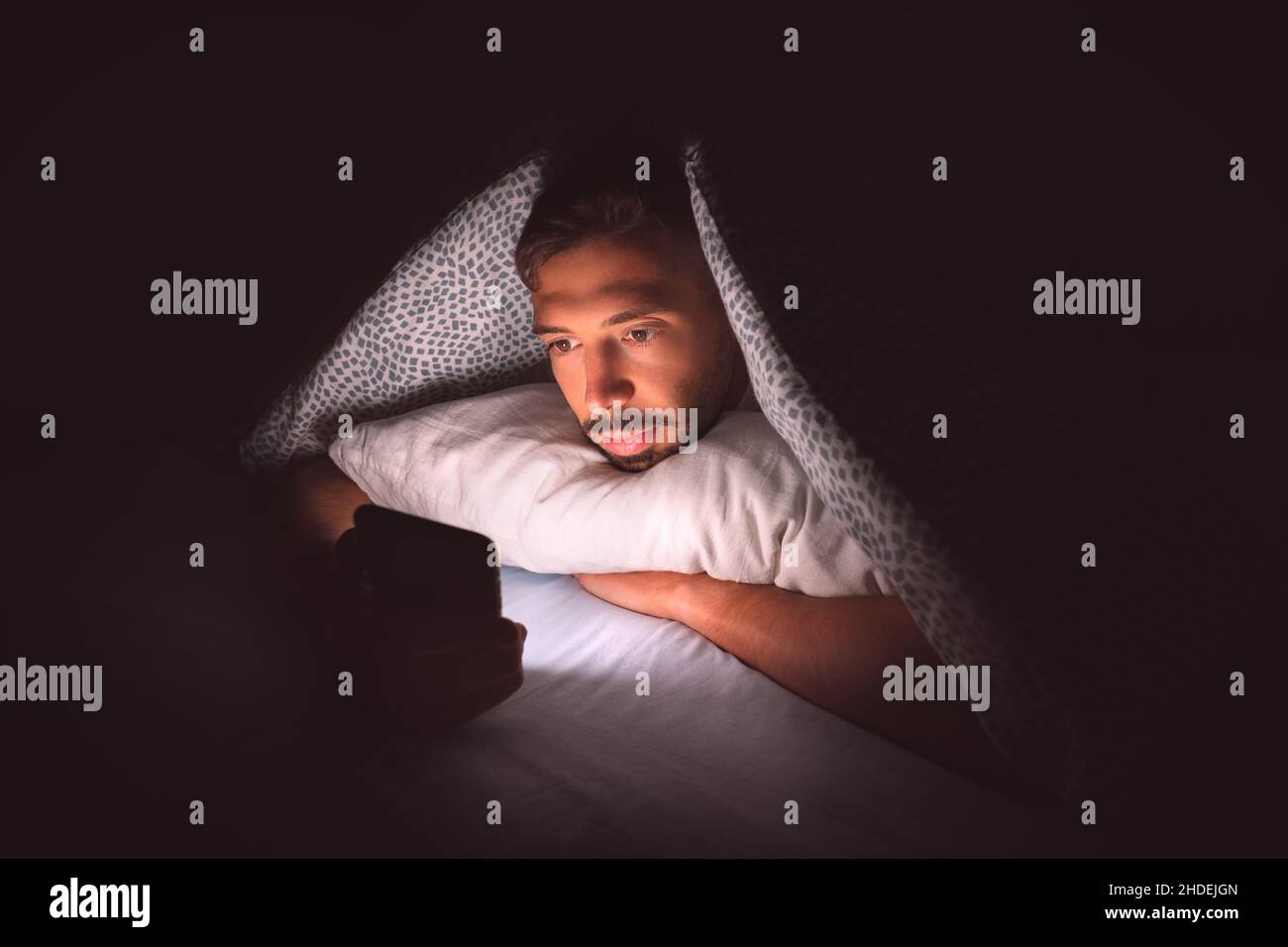 Jeune homme allongé dans un lit sous une couverture et utilisant un smartphone la nuit.Toxicomanie et communication dans les médias sociaux Banque D'Images
