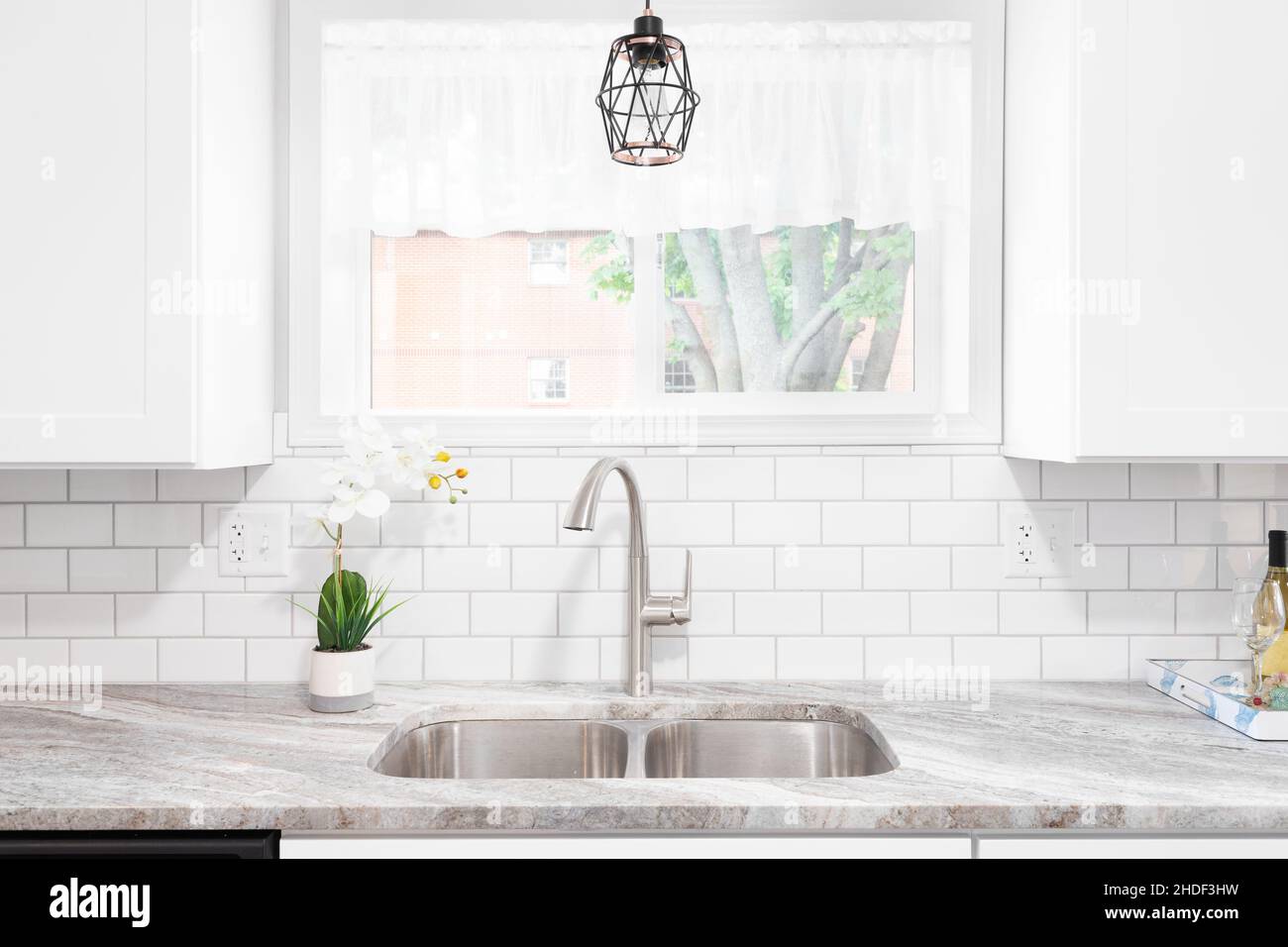 Le détail de l'évier de cuisine est réalisé avec un comptoir en marbre, des  armoires blanches, un robinet et un évier en acier inoxydable et une  lumière suspendue devant la fenêtre Photo