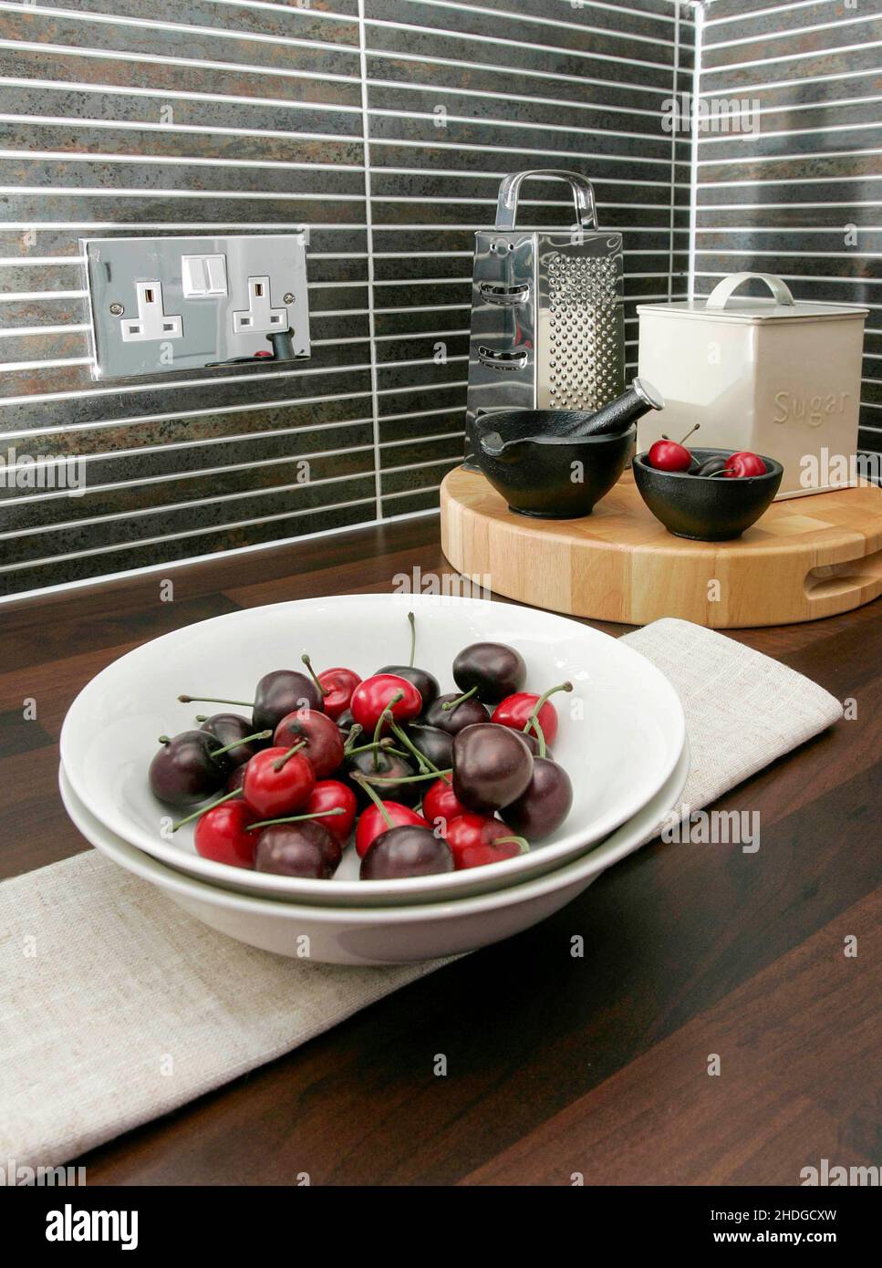 Bol de cerises sur le plan de travail de la cuisine, mortier pestel, sucrier, prise électrique chromée, carreaux mouchetés.Life est un bol de cerises Banque D'Images