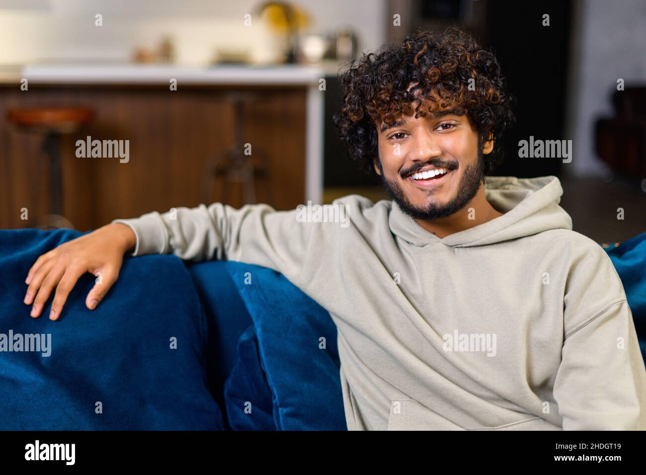 Portrait d'un jeune Indien insouciant assis sur le canapé et regardant la caméra, serein homme ethnique curly dans un pull à capuche décontracté dans une atmosphère détendue passer des vacances à la maison Banque D'Images
