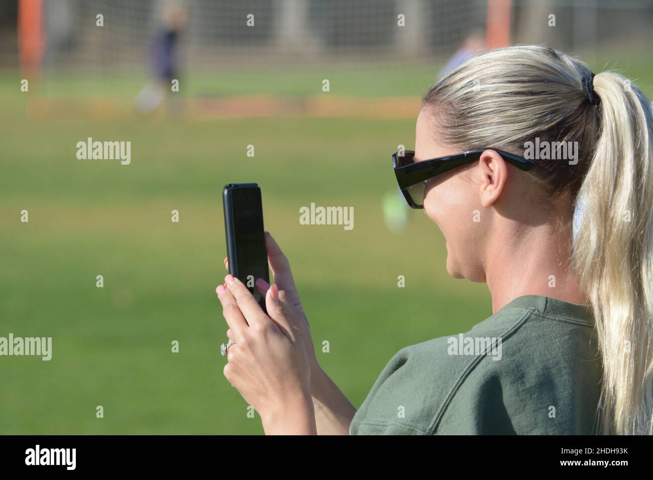 Jeune mère blonde utilisant le téléphone de vente pour enregistrer filles jeu de football, avec des lunettes de soleil et queue de cheval Banque D'Images