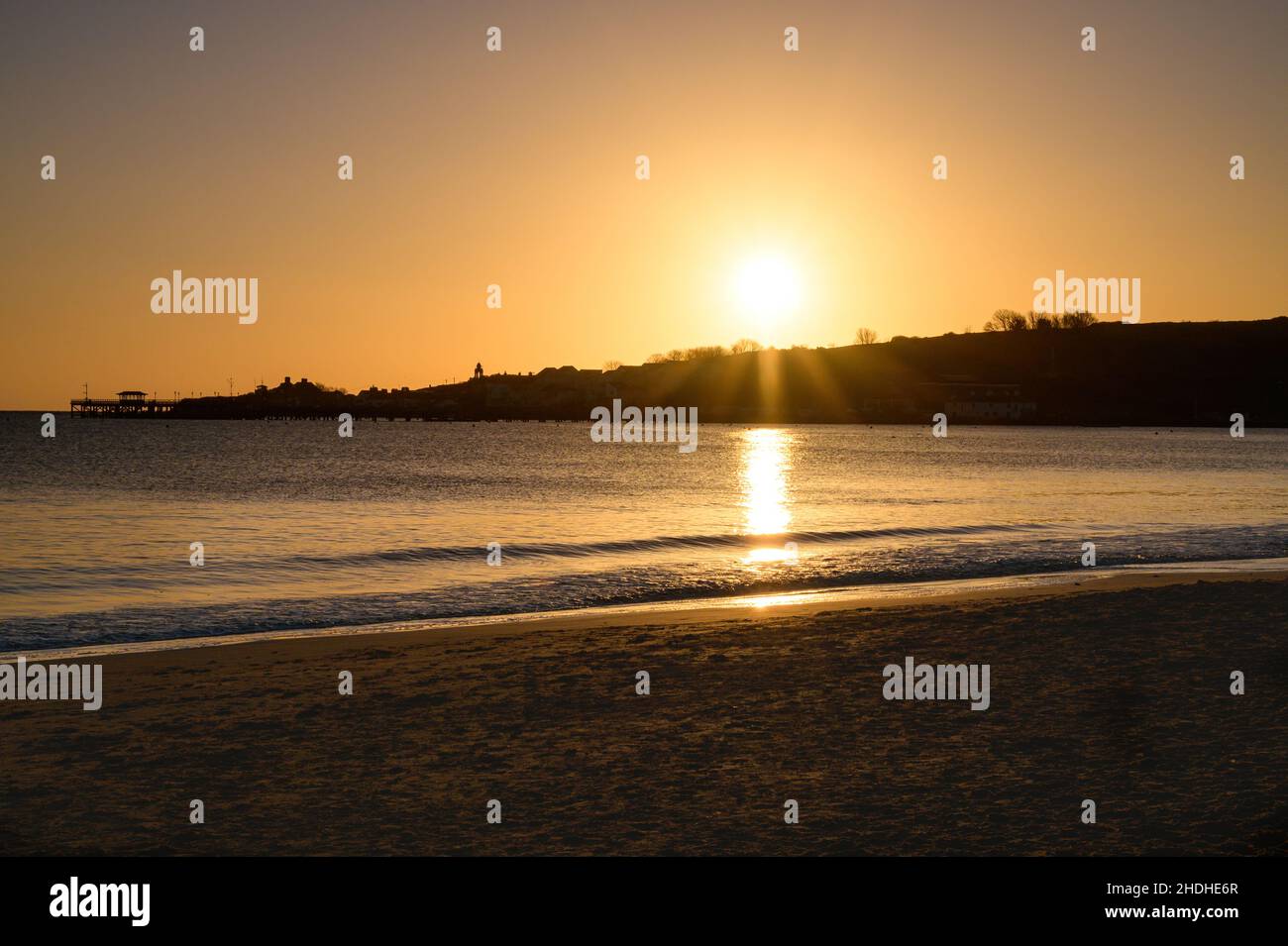 Lever du soleil à Swanage en regardant de l'autre côté de la baie avec la jetée de Swanage, le sommet des collines et la tour de l'horloge de Wellington en silhouette avec ciel clair et plage de sable. Banque D'Images