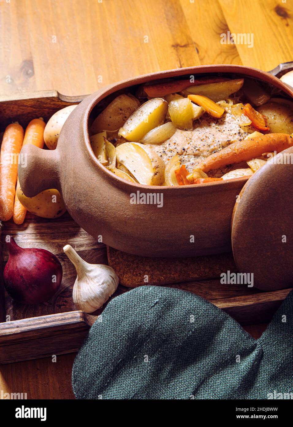 Vue latérale de la marmite en terre cuite avec rôti de porc lentement cuit et légumes à l'intérieur sur un plateau en bois et une table en bois, entouré de matières premières biologiques. Banque D'Images