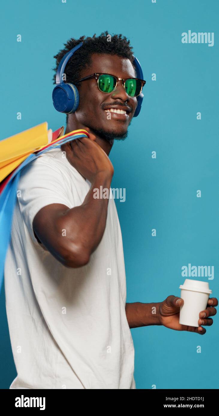 Homme heureux avec des sacs à main dansant après la frénésie de shopping. Personne joyeuse avec des lunettes de soleil écoutant de la musique sur les écouteurs tout en tenant des sacs avec des vêtements à vendre achat le vendredi noir Banque D'Images