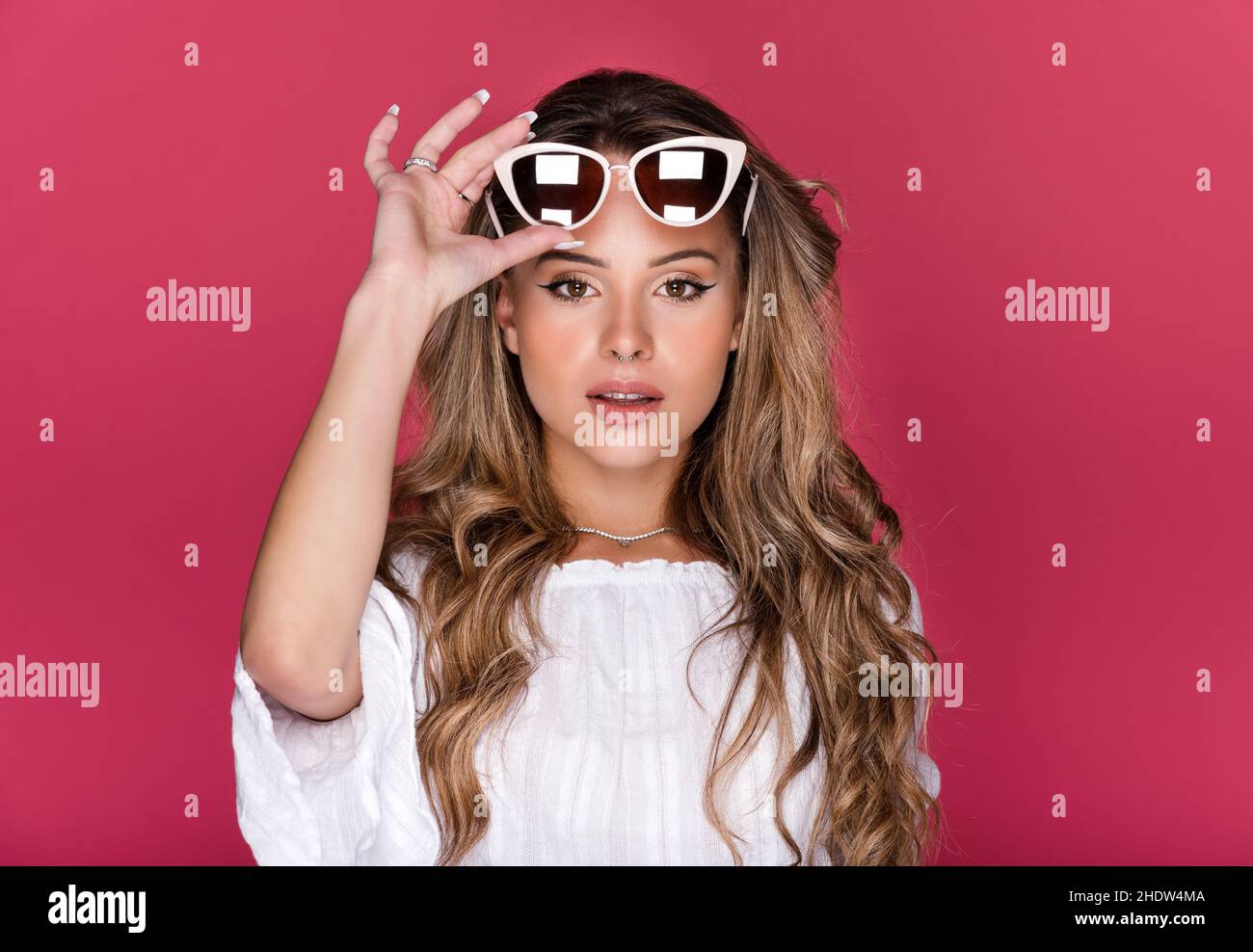 Jeune femme modèle avec de longs cheveux ondulés dans le chemisier portant des lunettes de soleil et regardant l'appareil photo contre fond rose dans le studio Banque D'Images