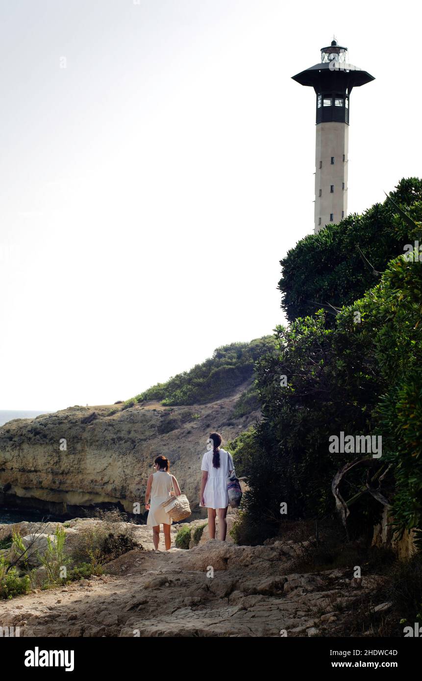 deux jeunes femmes méconnaissables avec des robes marchant vers le phare. touristes femmes explorent le chemin près de la falaise avec un grand sac chacun. Banque D'Images