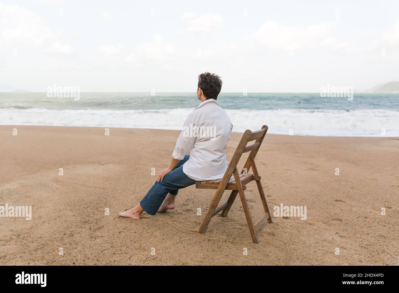 Heureuse femme à la retraite portant un châle assis sur une chaise, détendue sur le sable à la plage. Femme de race blanche Banque D'Images
