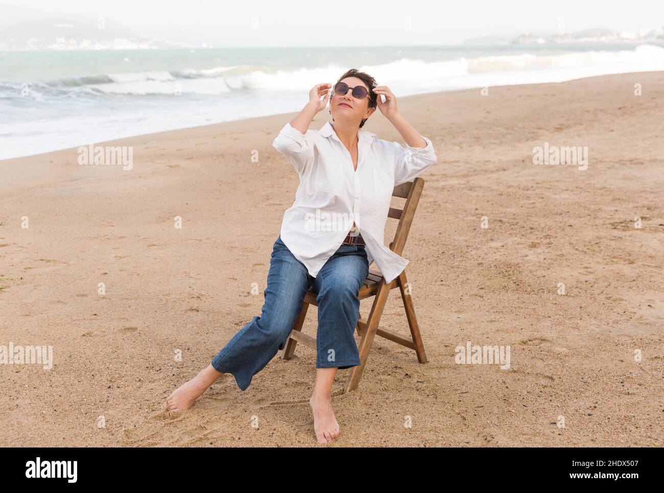 Heureuse femme à la retraite portant un châle assis sur une chaise, détendue sur le sable à la plage. Femme de race blanche Banque D'Images