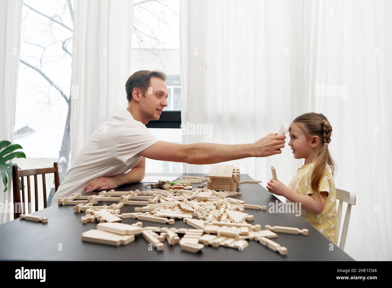 Vue latérale du papa vérifiant la température corporelle avec thermomètre infrarouge frontal de la fille assise à la table et jouant avec des blocs de bois Banque D'Images