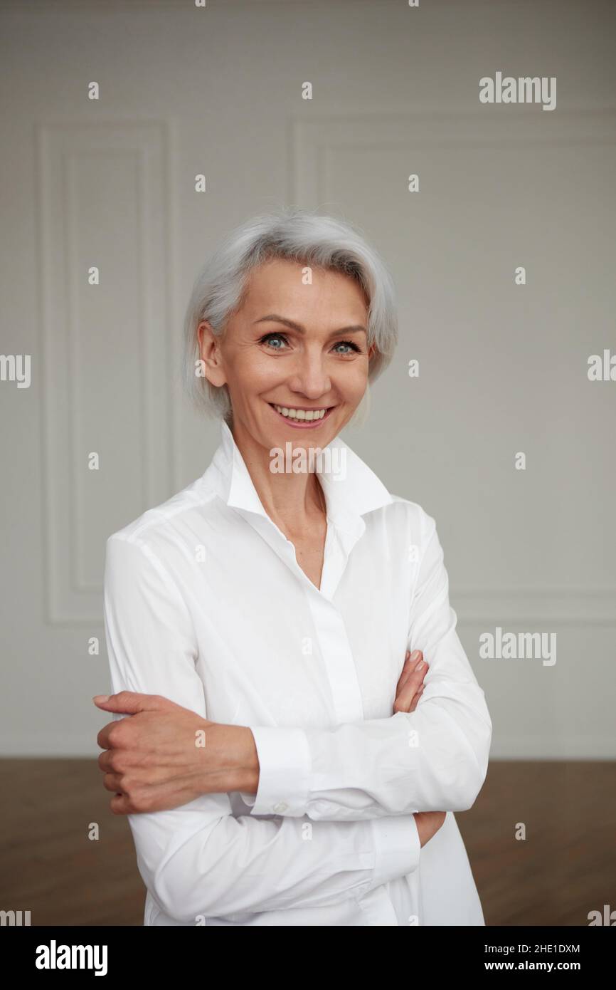 Portrait d'une femme adulte âgée et sûre d'elle-même avec des cheveux blancs regardant l'appareil photo dans une chemise blanche mains croisées mur d'arrière-plan Banque D'Images