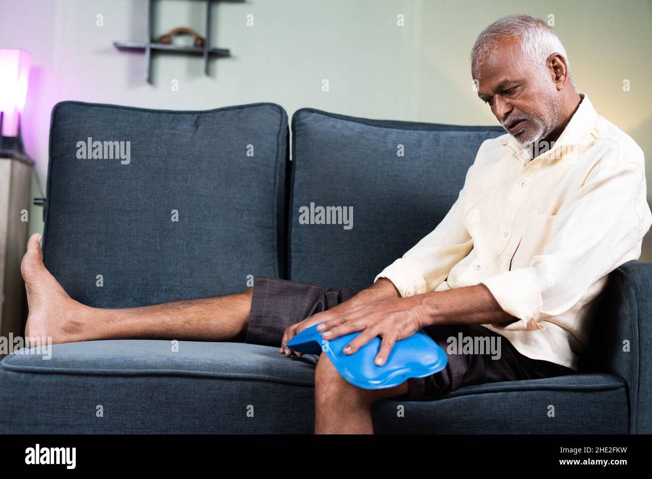 Homme senior utilisant un sac de massage d'eau chaude pour le soulagement de la douleur articulaire de genou tout en étant assis sur le canapé à la maison - concept de traitement d'eau chaude naturelle, muscle Banque D'Images