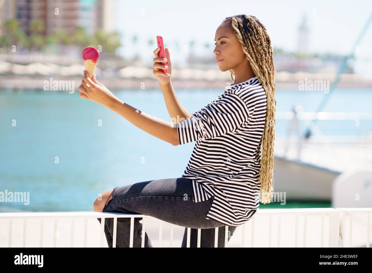 Fille noire photographiant avec son smartphone une crème glacée à la fraise qu'elle mange près de la mer Banque D'Images