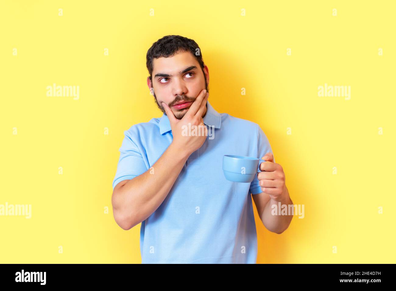 Jeune homme sérieux et pensif, tenant une tasse de café, isolé sur le mur jaune. Banque D'Images