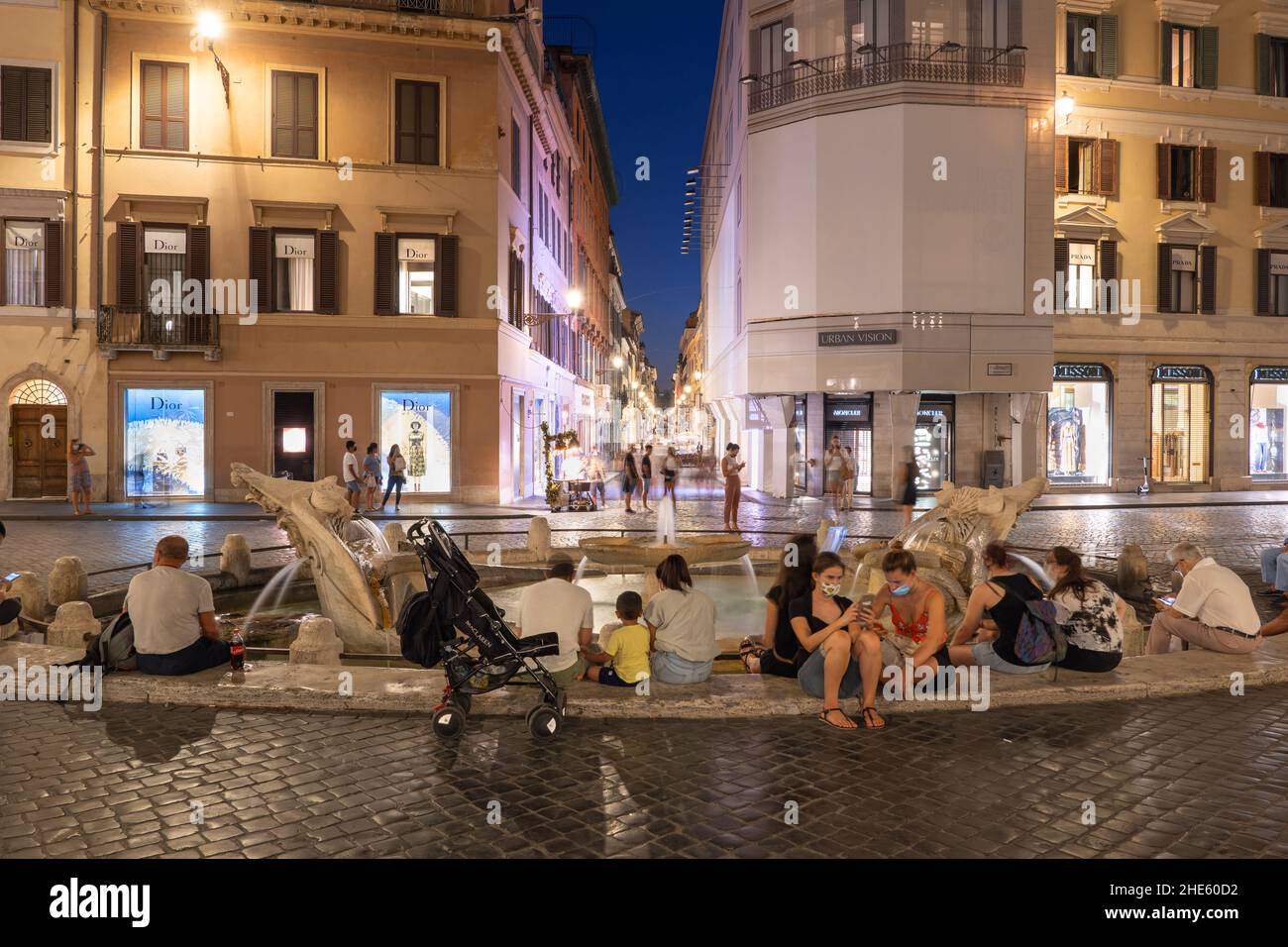 Italie, ville de Rome, les gens se détendent à la fontaine Barcaccia sur la Piazza di Spagna la nuit avec vue sur la rue via dei Condotti. Banque D'Images