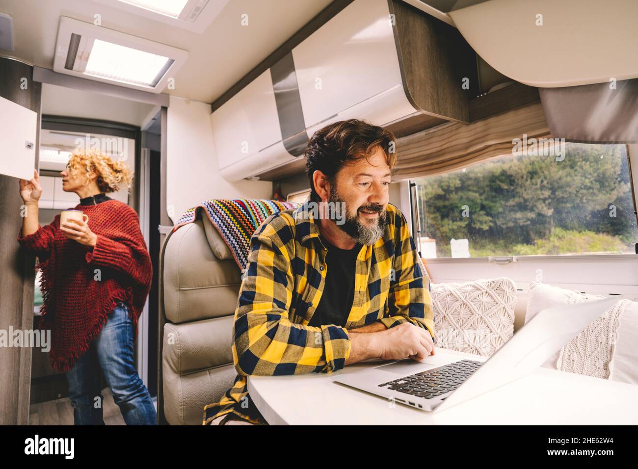 Les adultes voyagent en couple et apprécient le camping-car pour les vacances ou le style de vie vanlife - un homme heureux travaille sur un ordinateur portable sur la table et une femme lookin Banque D'Images