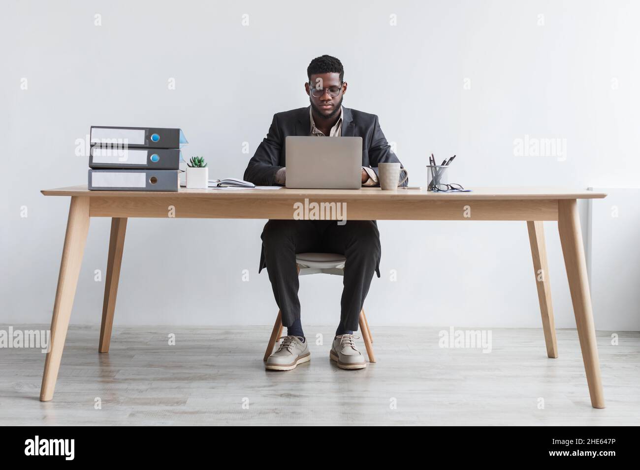 Pleine longueur de jeune homme noir concentré travaillant en ligne avec un ordinateur portable, assis au bureau contre le mur blanc du studio Banque D'Images