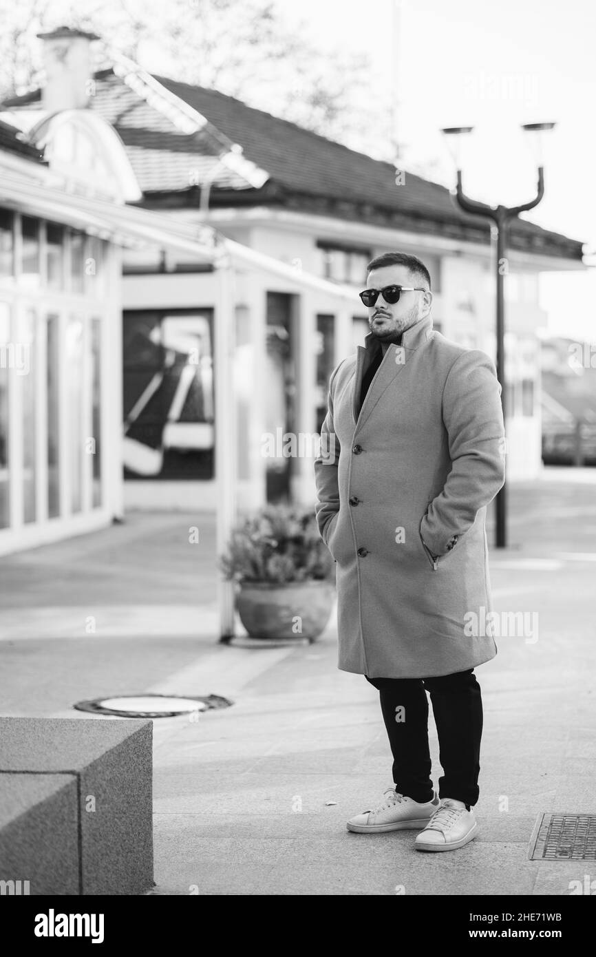 Un homme caucasien dans un manteau à la mode et des lunettes de soleil se tenant près d'un beau bâtiment Banque D'Images
